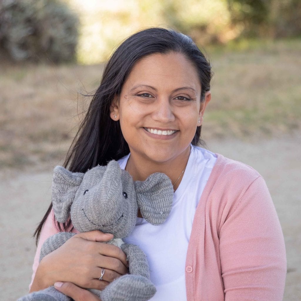 Miranda holding Adrian's elephant in a park in California. It is a sunny afternoon. Miranda is wearing a white shirt and pink cardigan and smiling. (Sarah Perry Photography)